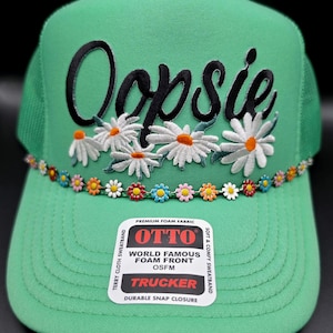 May include: A bright green trucker hat featuring the word Oopsie in black script embroidery above a cluster of white daisy appliques. A decorative trim of small, colorful enamel daisy charms lines the base of the front panel above the brim.