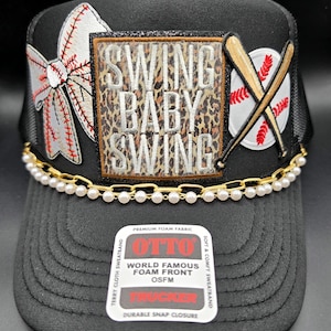 May include: Black trucker hat with a baseball theme. Features a white baseball bow, a leopard print patch with "SWING BABY SWING", and a baseball with crossed bats. A gold chain with pearls accents the brim. The hat has a snap closure.