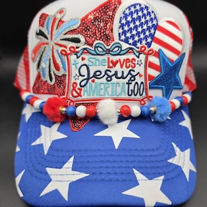 May include: A patriotic trucker hat with a white mesh back and a blue and white star-patterned brim. The front features red sequins, a blue star, and the text "She Loves Jesus & America too". Embellished with red, white, and blue beads and pom-poms.
