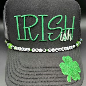 May include: Black trucker hat with the word "IRISHish" embroidered in green. A beaded band with the words "SHAKE YOUR SHAMROCKS" is across the front. A green shamrock is on the lower right side of the hat.
