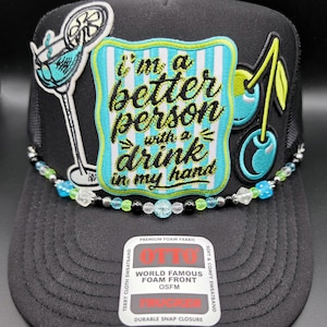 May include: A black trucker hat featuring a central patch with light blue and white stripes and the text "i'm a better person with a drink in my hand." The hat includes a cocktail glass patch, a cherry patch, and a beaded trim across the brim.