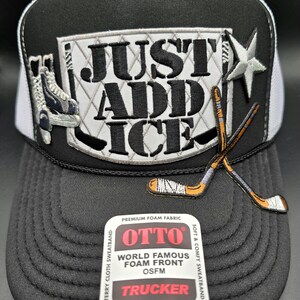 May include: Black and white trucker hat with the text "JUST ADD ICE" and hockey-themed embellishments. Features a star, ice skates, and crossed hockey sticks. The hat has a foam front and a durable snap closure.
