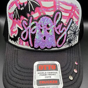 May include: A black and white trucker hat featuring a front panel decorated with a pink airbrushed spiderweb, a black bat, a spider, and a purple sequined ghost with the word "spooky" in script. The brim includes three small rhinestones.