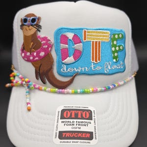 May include: White trucker hat with a colorful beaded string and a patch featuring an otter wearing sunglasses and a pink polka-dot floatie. The patch reads "DTF down to float". The hat has a foam front and a snap closure.