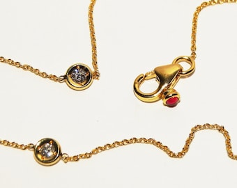 May include: A gold chain necklace featuring multiple bezel-set clear round stones spaced along the length of the chain. The necklace includes a gold lobster claw clasp with a small, round, red accent stone attached near the closure mechanism.