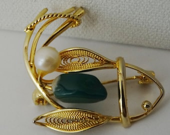 Puede incluir: Un broche de filigrana dorada con una piedra verde y una perla blanca. La broche tiene la forma de una hoja estilizada.