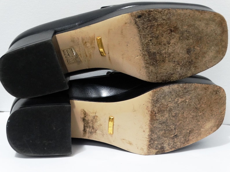 May include: Black leather loafers with a gold logo on the sole. The shoes have a chunky heel and a square toe.