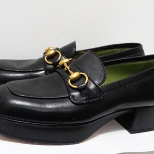 May include: Black leather platform loafers with gold-tone hardware. The loafers have a chunky platform sole and a square toe.