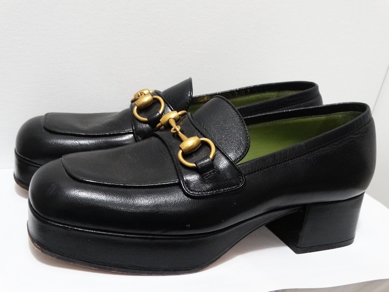 May include: Black leather platform loafers with gold-tone hardware. The loafers have a chunky platform sole and a square toe.