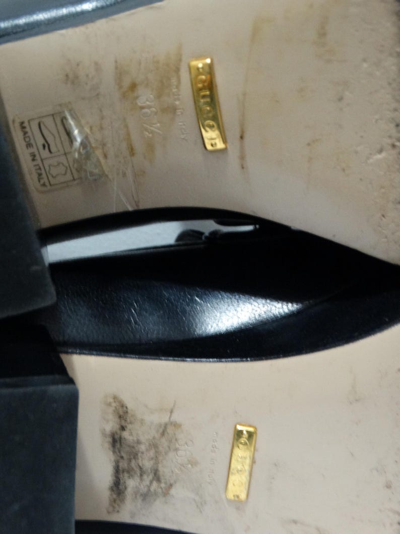 May include: Close-up of the bottom of a pair of black leather shoes. The shoes have a gold label that reads "Gucci" and "Made in Italy".