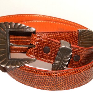 May include: A brown leather belt with a silver buckle featuring a shell design. The belt is made of a textured leather that resembles snakeskin.