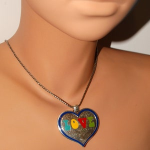 May include: A silver heart-shaped pendant necklace with a blue border and the word "LOVE" spelled out in colorful enamel. The letters are in a whimsical font and each letter is a different color.