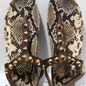 Könnte beinhalten: Braune Ledersandalen mit goldenen Nieten und Schlangenhautmuster.