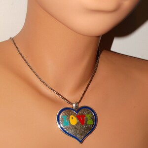 May include: A silver heart-shaped pendant necklace with a blue border and the word "LOVE" spelled out in colorful enamel. The letters are outlined in silver and the background of the heart is filled with a silver swirl pattern.