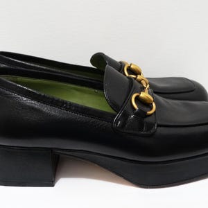 May include: Black leather platform loafers with gold hardware. The loafers have a chunky heel and a rounded toe.