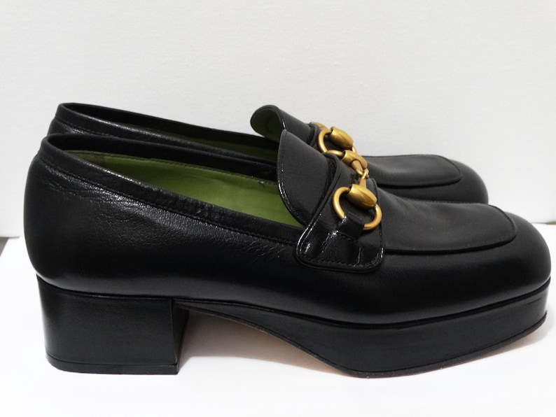 May include: Black leather platform loafers with gold hardware. The loafers have a chunky heel and a rounded toe.