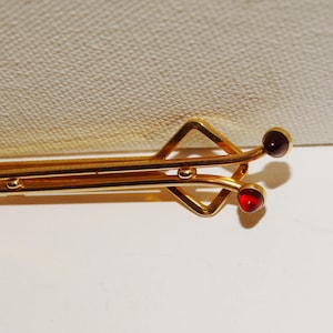 May include: A gold-toned tie clip with a geometric design. The clip features a diamond shape and two red, round gemstones. The clip is designed to hold a tie in place and adds a touch of elegance.