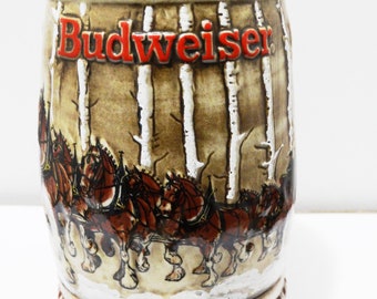 Könnte beinhalten: Ein Budweiser Bierkrug aus Keramik mit einer braunen und weißen Winterszene mit einem Team von Clydesdale-Pferden, die einen Wagen ziehen. Der Krug hat einen roten und weißen Rand und das Budweiser-Logo ist rot.