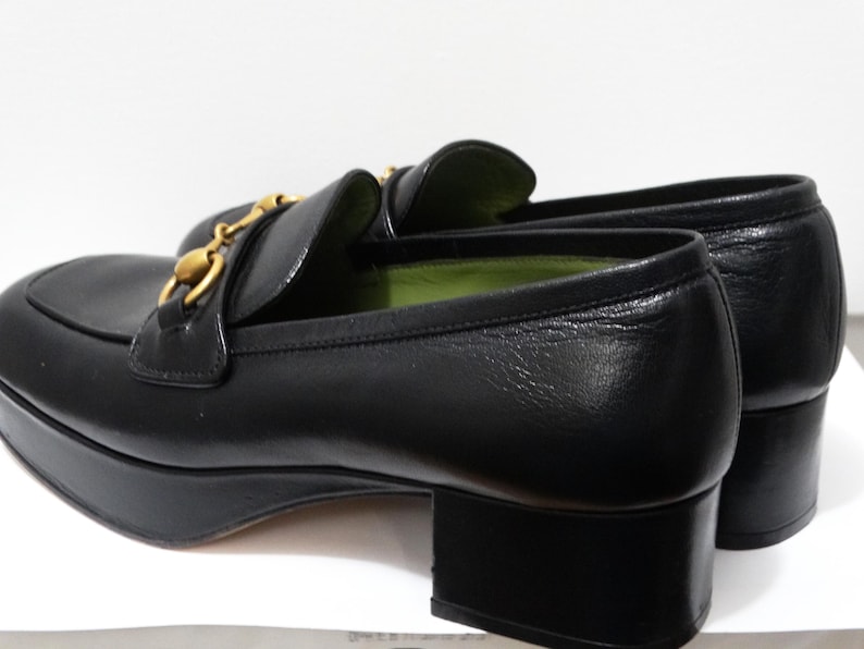May include: Black leather platform loafers with a gold chain detail. The loafers have a green lining and a chunky heel.