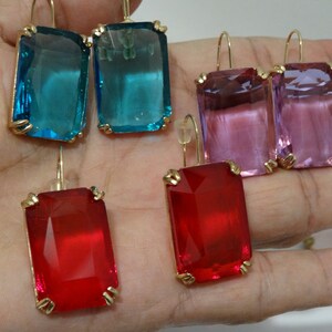 May include: Gold-toned rectangular drop earrings with large, colorful, faceted gems. The earrings are available in teal, pink, and red. The gems are set in gold-colored metal with a simple hook closure.