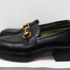 May include: Black leather platform loafers with gold chain detail. The loafers have a chunky heel and a square toe.