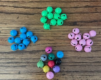 May include: An assortment of jingle bells in various colours, including green, blue, pink, red, black, yellow, and grey. The bells are arranged in groups on a wooden surface.