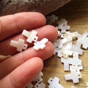 May include: A collection of white mother of pearl cross charms with a hole for stringing. The charms are scattered on a wooden surface and a hand is holding a few of the charms.