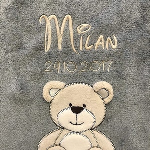 Può includere: Una coperta di peluche grigia con un orsacchiotto di peluche beige ricamato e il testo "Milan 24.10.2017".