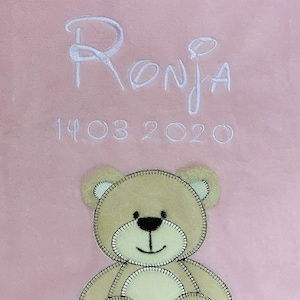 May include: A pink baby blanket with a white embroidered name "Ronja" and the date "14.03.2020". A brown teddy bear with white accents is sewn on the blanket.