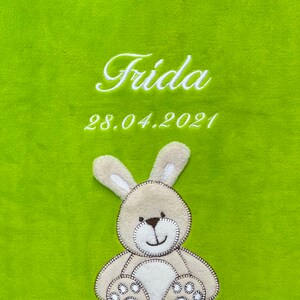 Könnte beinhalten: Eine grüne Plüschdecke mit dem Namen "Frida" und dem Datum "28.04.2021" in weiß gestickt. Ein beigefarbener Plüschhase mit braunen Nähten ist auf die Decke genäht.