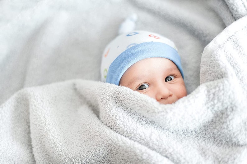 Puede incluir: Un beb&eacute; con un gorro azul y blanco con dise&ntilde;os coloridos asoma desde una manta blanca.