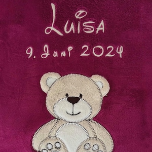 Può includere: Una coperta di peluche rosa con un orsacchiotto di peluche ricamato in bianco e il testo "Luisa 9. Jan 2024".
