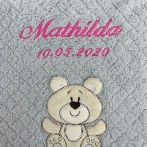 May include: A gray plush blanket with a pink embroidered name "Mathilda" and the date "10.05.2020". A beige embroidered teddy bear is in the center of the blanket.