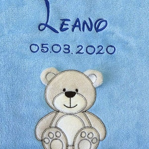 Pode incluir: Um cobertor de bebê azul claro com um aplique de urso de pelúcia branco. O cobertor é bordado com o nome "Leano" e a data "05.03.2020".