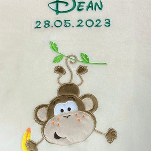 Könnte beinhalten: Cremefarbene Decke mit einem gestickten Affen, der eine Banane hält. Der Affe ist braun, beige und weiß mit orangefarbenen Akzenten. Der Name "Dean" und das Datum "28.05.2023" sind grün gestickt.