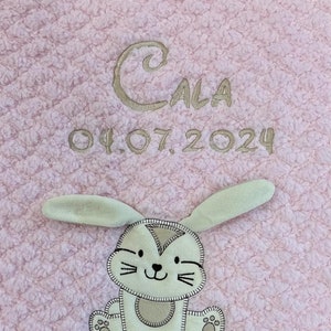 Può includere: Una copertina rosa per neonati con un'applicazione a forma di coniglio e il nome "CALA" ricamato, insieme alla data "04.07.2024".