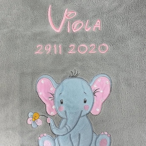 Op de afbeelding: Een grijze pluche deken met roze geborduurde tekst die "Viola 2911 2020" luidt. Een blauwe geborduurde olifant met roze oren en een gele bloem bevindt zich in het midden onderaan de deken.