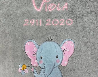 Puede incluir: Una manta de felpa gris con texto bordado en rosa que dice "Viola 2911 2020". Un elefante bordado azul con orejas rosas y una flor amarilla se encuentra en el centro inferior de la manta.