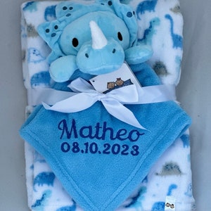 Könnte beinhalten: Eine blaue und weiße Babydecke mit einem Dinosaurier-Druck. Die Decke ist mit dem Namen "Matheo" und dem Datum "08.10.2023" personalisiert. Sie wird mit einem passenden blauen Dinosaurier-Plüschtier geliefert.