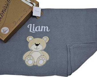 以下が含まれることがあります： 刺繍されたベージュのテディベアと白い筆記体でLiamと書かれたグレーのニットブランケット。ブランケットは、白いリボンとCotonissimoのタグ、綿のアイコンが付いた茶色の段ボール製ギフトボックスの隣に配置されています。