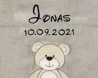 Puede incluir: Una manta de bebé beige con el nombre "Jonas" bordado en negro sobre la fecha "10.09.2021". Una aplicación de oso de peluche crema con costuras marrones está centrada en la parte inferior.