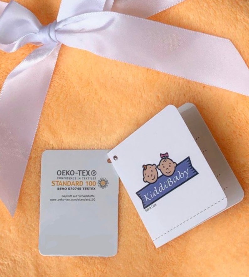 Puede incluir: Una manta de beb&eacute; de color melocot&oacute;n con una cinta de sat&eacute;n blanca atada alrededor. La manta tiene dos etiquetas adjuntas. Una etiqueta es blanca con el texto "OEKO-TEX&reg; CONFIDENCE IN TEXTILES STANDARD 100 BEHO 070745 TESTEX Gepr&uuml;ft auf Schadstoffe www.oeko-tex.com/standard100" y la otra etiqueta es blanca con el texto "KiddiBaby Safe & Care" y una imagen de dibujos animados de dos beb&eacute;s.