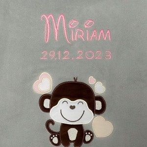Könnte beinhalten: Eine graue Babydecke mit rosa Stickerei, auf der "Miriam" und "29.12.2023" steht. Ein brauner Affe mit einem weißen Lächeln und rosa Herzen ist auf die Decke gestickt.
