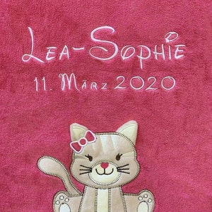 Op de afbeelding: Een roze babydeken met witte geborduurde tekst "Lea-Sophie 11. März 2020" en een bruine en witte geborduurde kat met een roze strik.