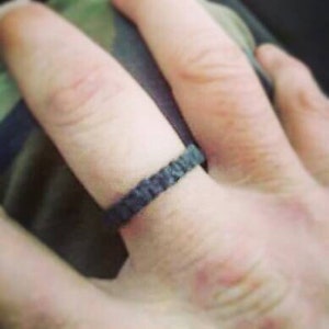 May include: A dark gray ring with a textured, uneven surface. The band has a matte finish and a consistent width, encircling the finger. The material appears to have a rough, hammered, or organic appearance, creating a distinct tactile look.