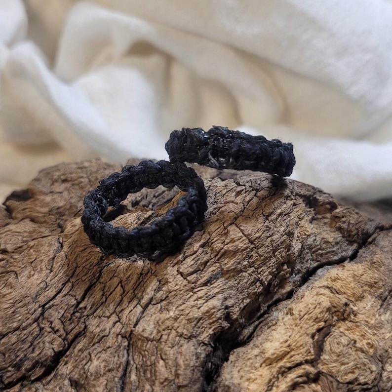 May include: Two black rings crafted with a textured, knotted cord design. The rings feature a continuous, woven pattern throughout the band, creating a rugged and tactile surface. The dark, matte finish of the cord contrasts with the intricate knots.