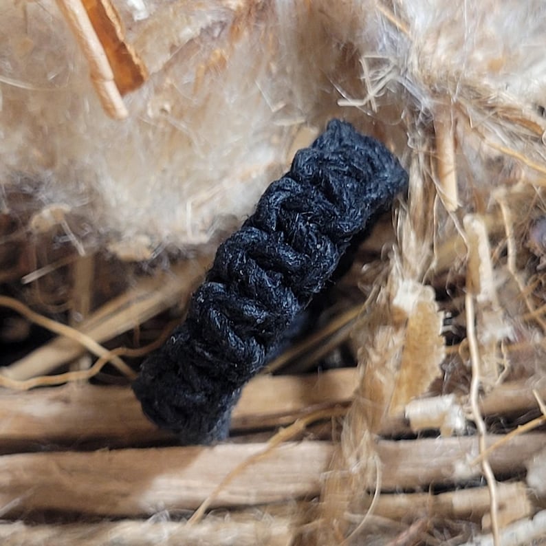 May include: A black ring constructed from tightly woven cord. The band features a distinct, repeating knotted pattern throughout its circumference, creating a textured, cylindrical surface with a matte finish and a consistent dark black color.