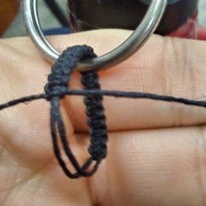 May include: A black macrame cord being knotted around a silver-toned metal ring. The cord is thin and dark, showing a series of tight, uniform knots forming a loop structure. The process captures the intricate weaving technique of the fiber.