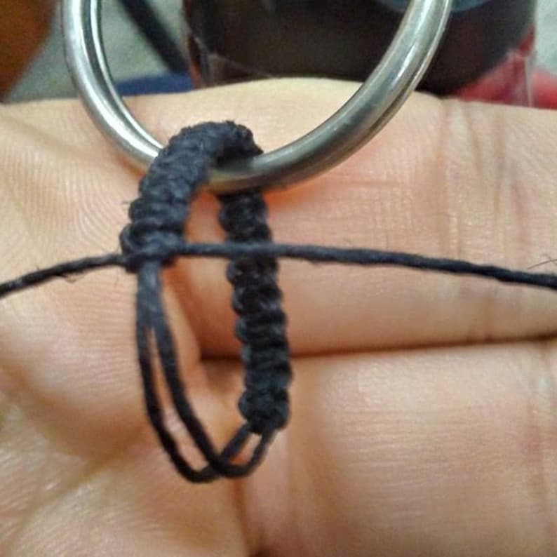 May include: A black macrame cord being knotted around a silver-toned metal ring. The cord is thin and dark, showing a series of tight, uniform knots forming a loop structure. The process captures the intricate weaving technique of the fiber.