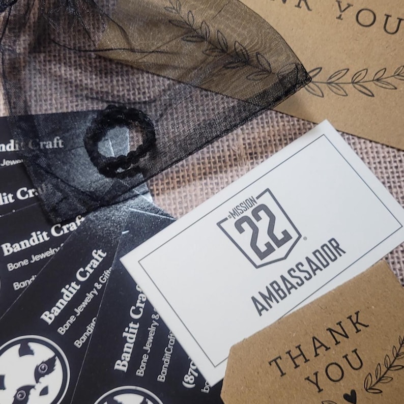 May include: A black ring sits on a sheer black organza pouch next to several black Bandit Craft business cards and a white Mission 22 Ambassador card. Two brown kraft paper cards with the text Thank You and leaf illustrations are also visible.